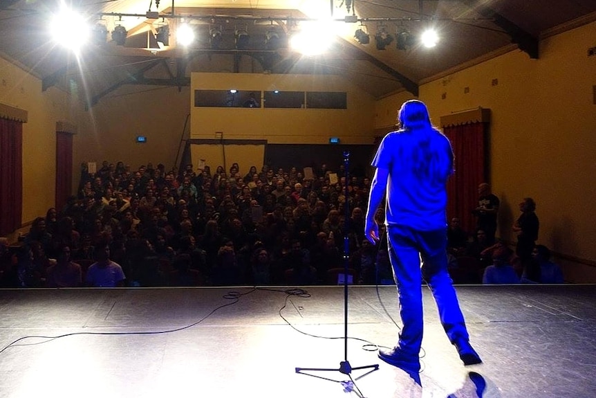 A man lit up in blue stands on a stage in front of an audience