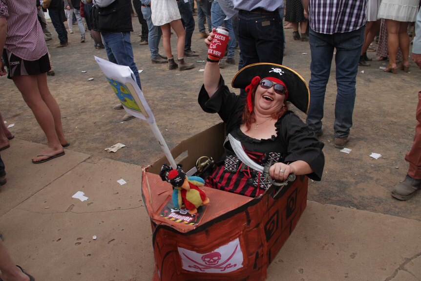 A woman wearing a tricornered hat emblazoned with the Jolly Roger hoists a tinnie skyward while sitting in a cardboard boat.