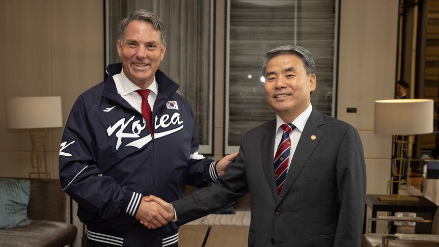Marles, wearing a sport jacket labelled 'Korea', shakes hands with a man in a suit, both smiling at the camera.