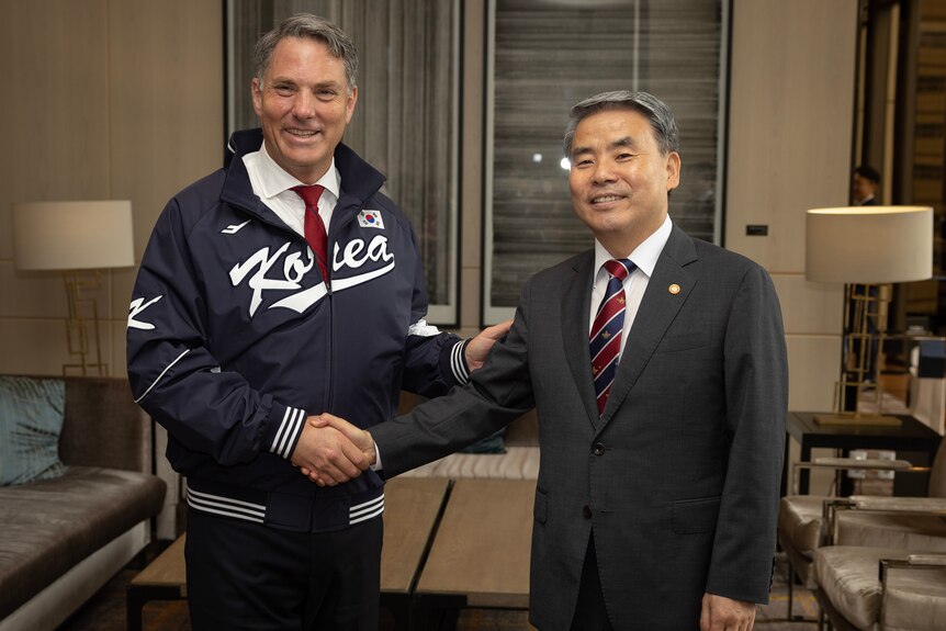 Marles, wearing a sport jacket labelled 'Korea', shakes hands with a man in a suit, both smiling at the camera.