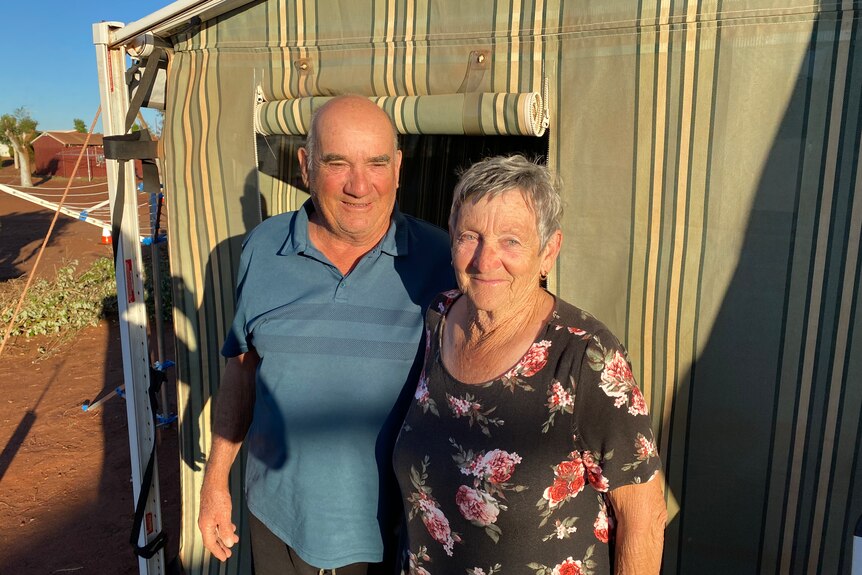 A bald man with blue polo shirt and a short-haired woman in floral dress stand in front of a caravan annex