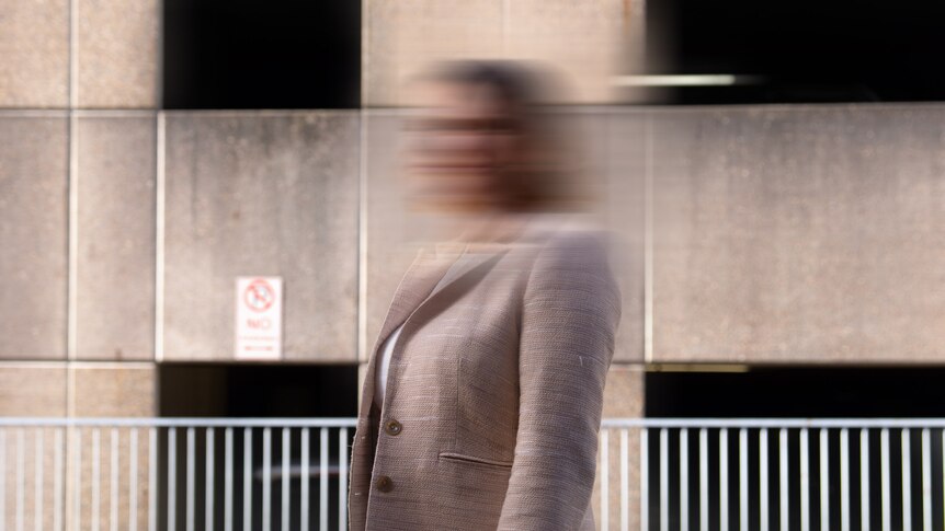 An unidentifiable woman in a creme blazer stands in front of a concrete building
