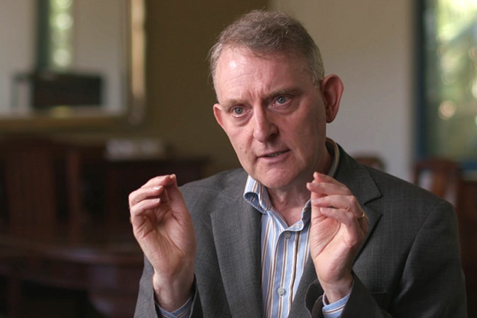 A man in a grey jacket and striped blue shirt gestures with his hands.