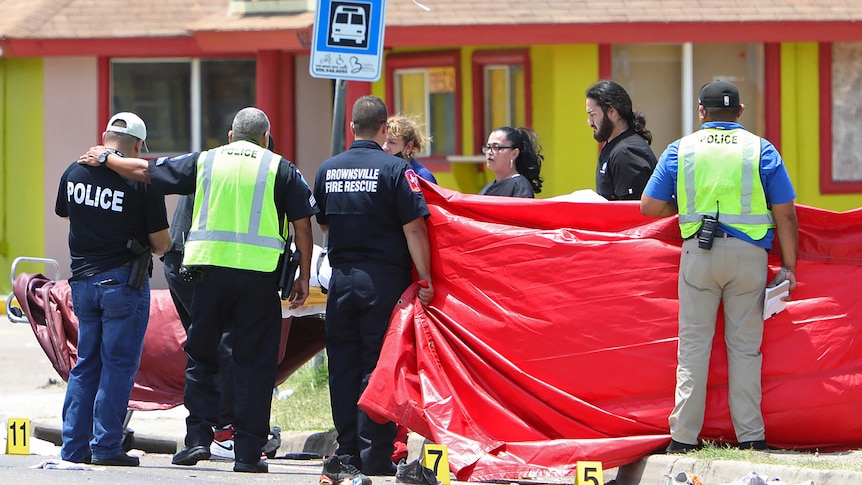 Emergency workers stand in front of a stretcher with large red plastic sheet blocking view as shoes lay, scattered, on road.