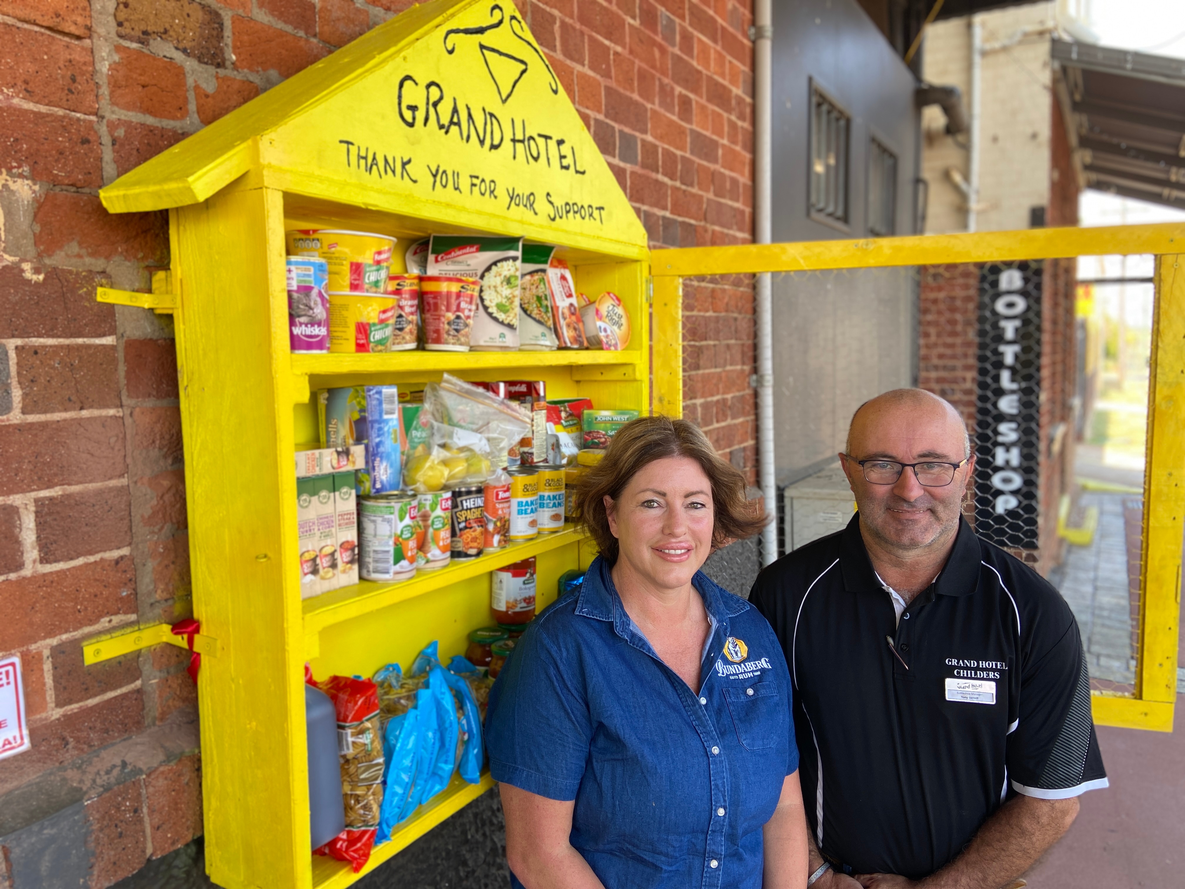 A woman and a man leaning against a bright yellow box filled with food attached to the outside of a building