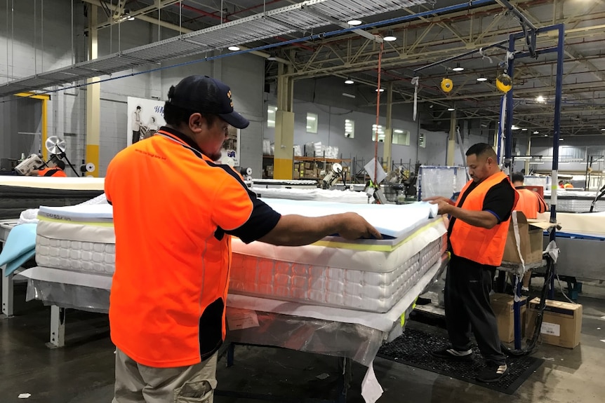 Two workers building a mattress at a factory, wearing orange, high visibility vests.