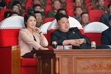 Kim Jong Un sits in a crowded theatre next to a woman in a cream and black suit