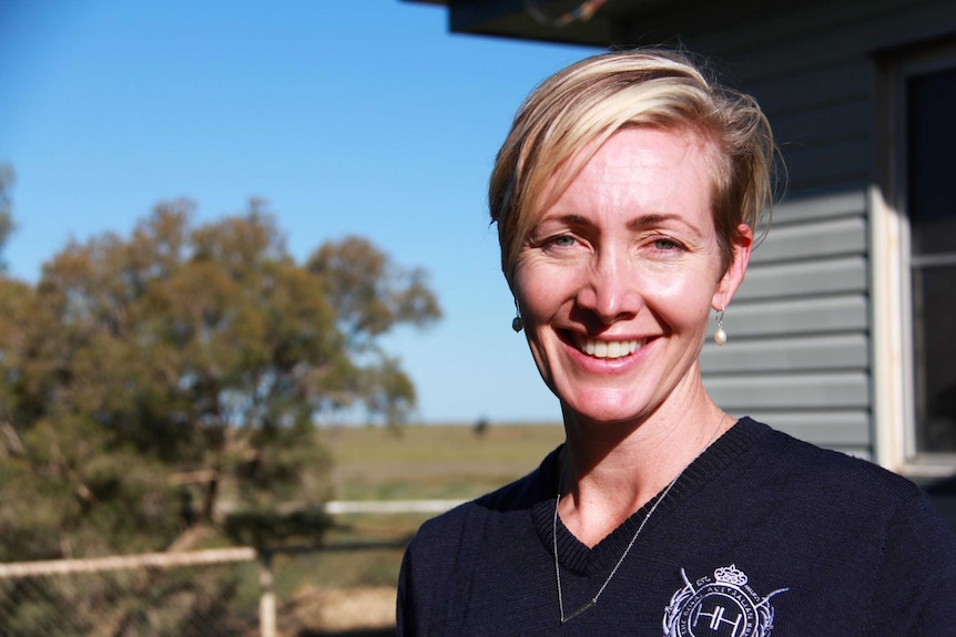 A close up of a woman with short blonde hair in front of a country backdrop.