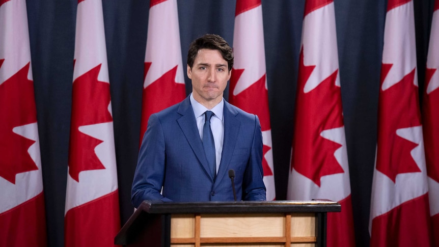 Prime Minister Justin Trudeau stands wearing a blue suit and tie with seven Canadian flags in the background.