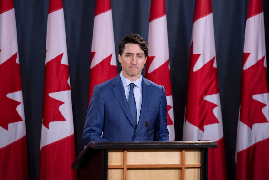 Prime Minister Justin Trudeau stands wearing a blue suit and tie with seven Canadian flags in the background.