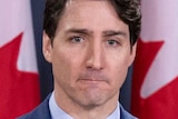 Prime Minister Justin Trudeau stands wearing a blue suit and tie with seven Canadian flags in the background.