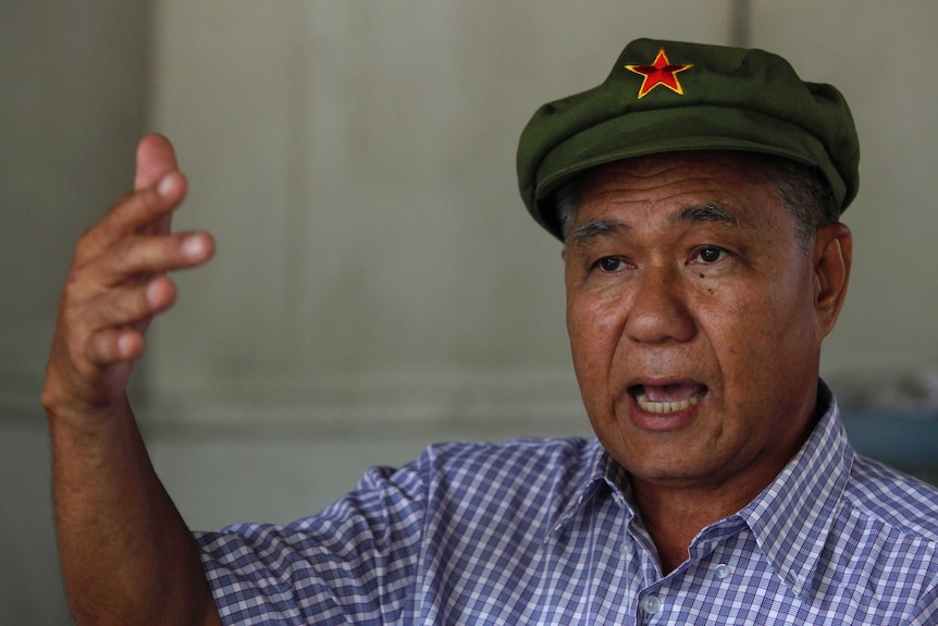 An older man gesticulating while wearing a green cap bearing a red star