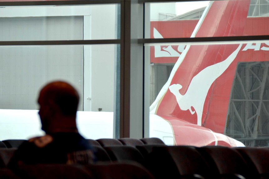 A man sits in a departures lounge at Sydney Airport, with a Qantas plane at the gate