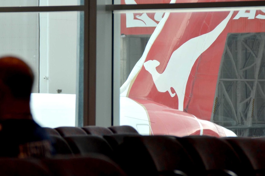 A silouette of a man sitting inside an airport terminal as a Qantas plane, with a red wing and white kangaroo, moves past