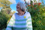 A grey haired woman looking to the side and smiling. Blue sky and green shrubs behind her.
