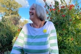 A grey haired woman looking to the side and smiling. Blue sky and green shrubs behind her.