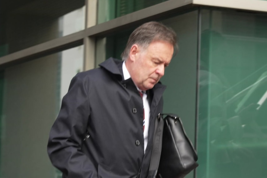 A middle aged man in a black coat looks at the ground with a serious expression. 