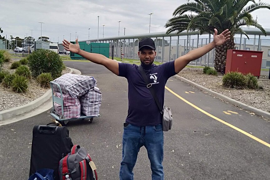 A man with his arms outstretched, with several bags of belongings to his right.