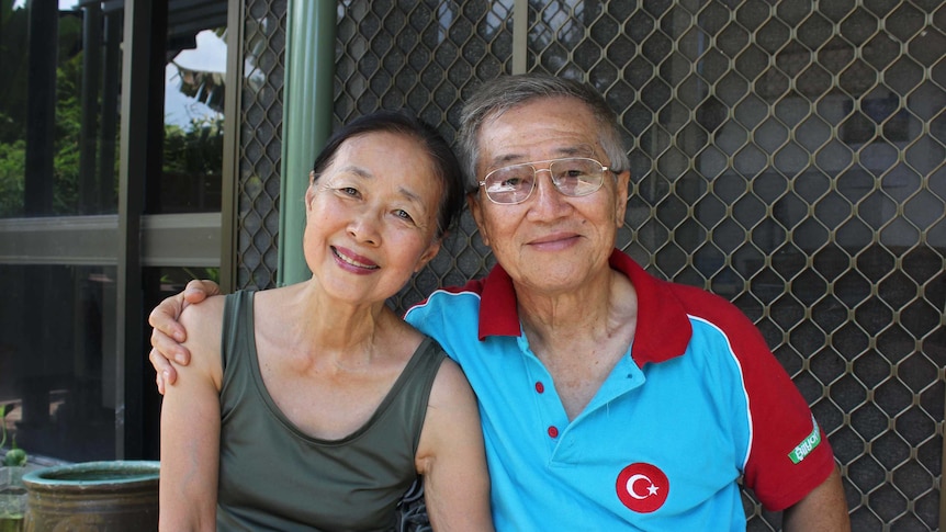 A elderly Malaysian couple sit with their arms around each other while smiling.