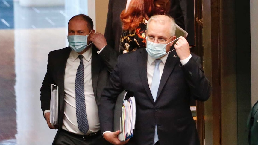 Two men in suits and wearing face masks walk through a door into a green room.