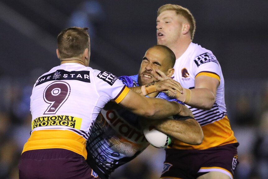 A Cronulla NRL player holds the ball as he is tackled by two Brisbane opponents.