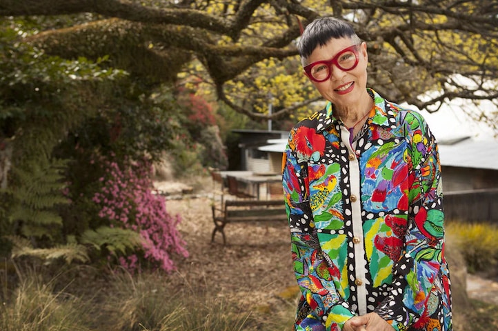 An older woman with short, dark hair, stylish spectacles and a brightly coloured shirt, standing outside and smiling.