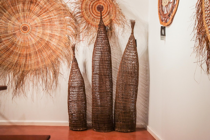 Three An-gujechiya (fish traps) made from jungle vine stand tall in a gallery space in front of wall weavings.