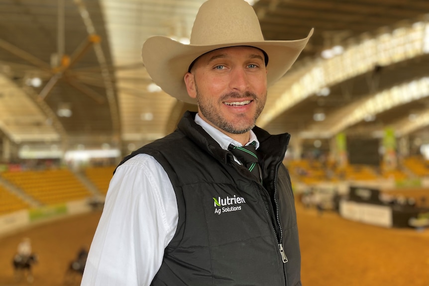 A man in a hat and black vest stands at the edge of big competition ring.