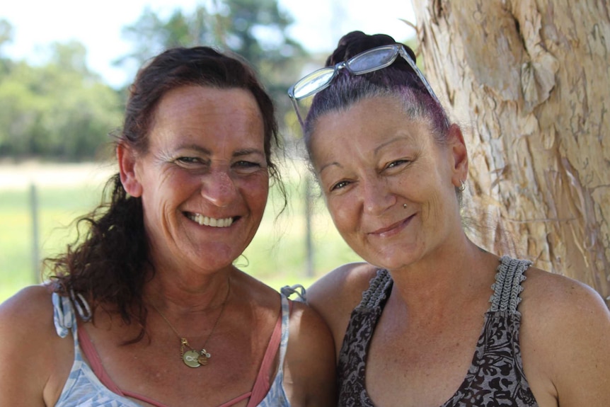 Two women, one has a arm around the other's shoulder, standing outside under a tree on a hot sunny day and smiling