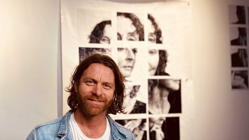Photographer Toby Burrows wearing a denim jacket and white t shirt standing in front of one of his images in an art gallery