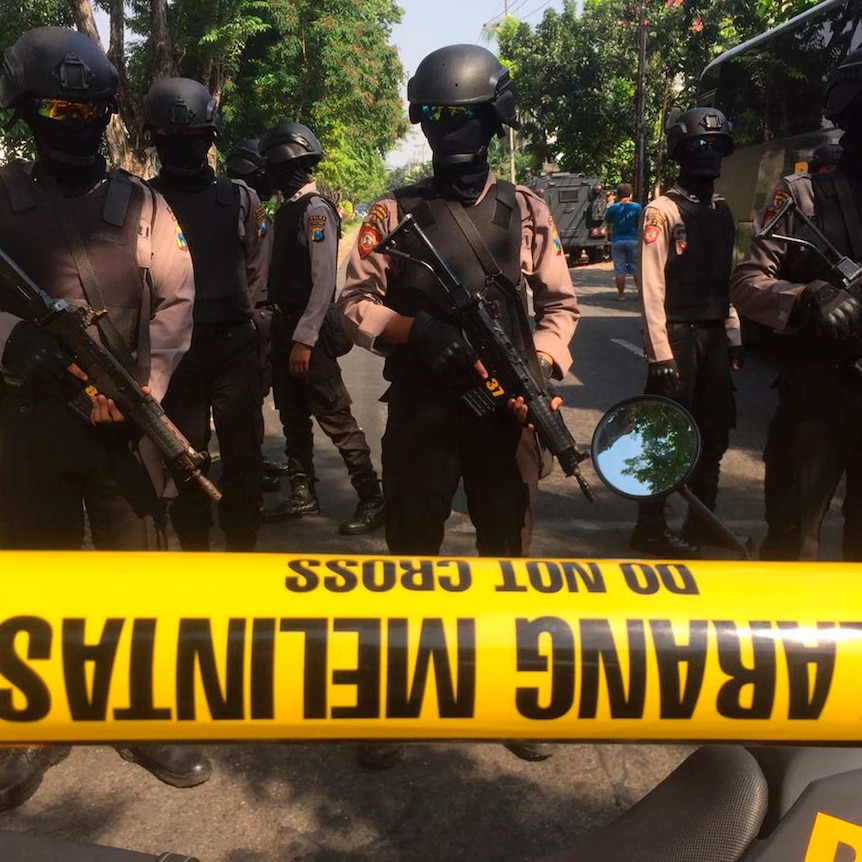 Police in helmets, goggles, and their faces covered, hold large guns as they stand guard behind a yellow tape on a road.