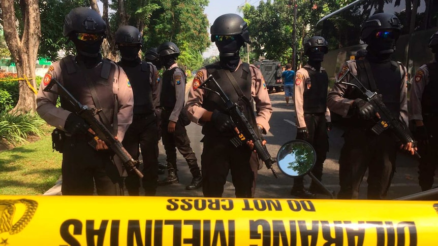 Police in helmets, goggles, and their faces covered, hold large guns as they stand guard behind a yellow tape on a road.