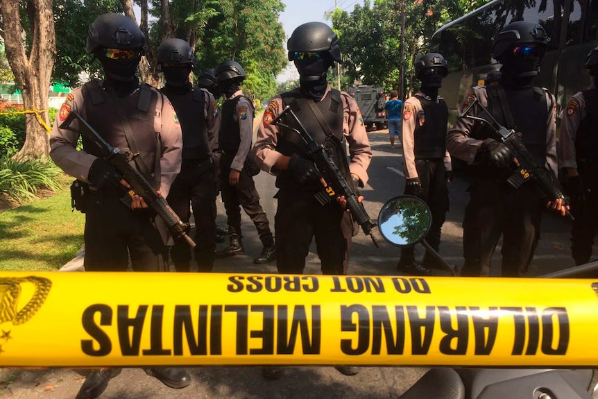 Police in helmets, goggles, and their faces covered, hold large guns as they stand guard behind a yellow tape on a road.