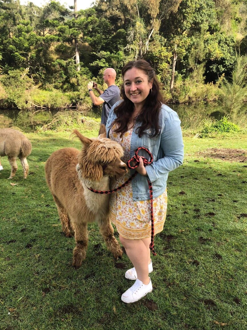 A photo of Jess smiling at the camera while standing next to an alpaca. She's wearing a denim jacket and floral dress.