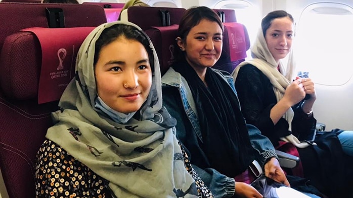 Three young women sit in a row in aeroplane seats. Two are wearing head scarves.