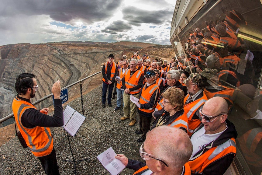 A group of people dressed in orange, hi-vis vests sing on the edge of the Super Pit gold mine.
