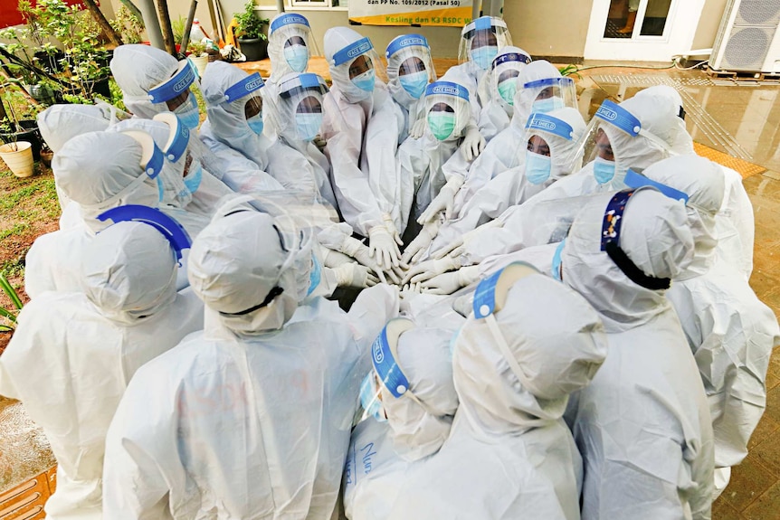 A group of people in white PPE with shields and masks on and heads covered put their hands in.