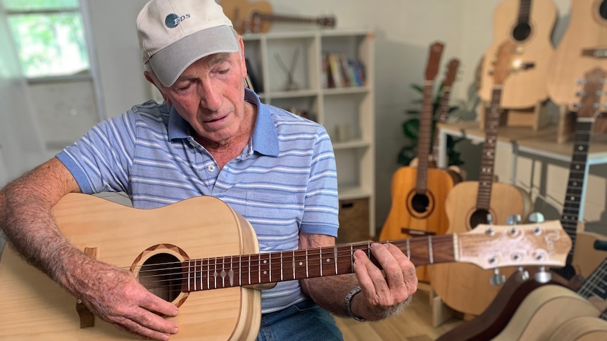 An older man wearing shorts and a striped t-shirt sits on a chair and plays a handmade guitar.