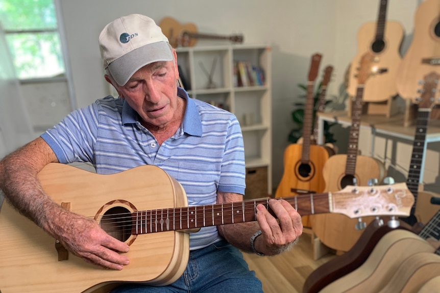 An older man wearing shorts and a striped t-shirt sits on a chair and plays a handmade guitar.