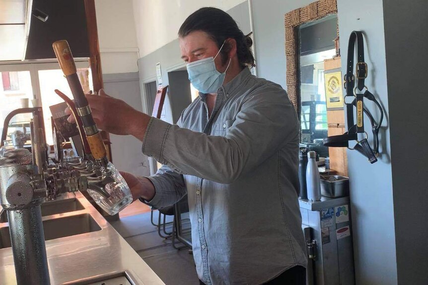 A man wearing a blue face mask stands behind a bar, pulling a beer.
