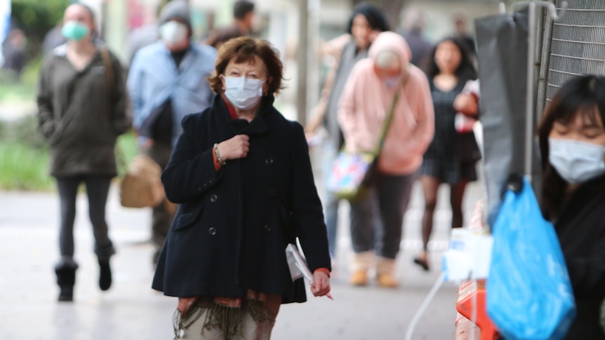 Peopole of all ages are walking outdoors, wearing blue surgical masks.