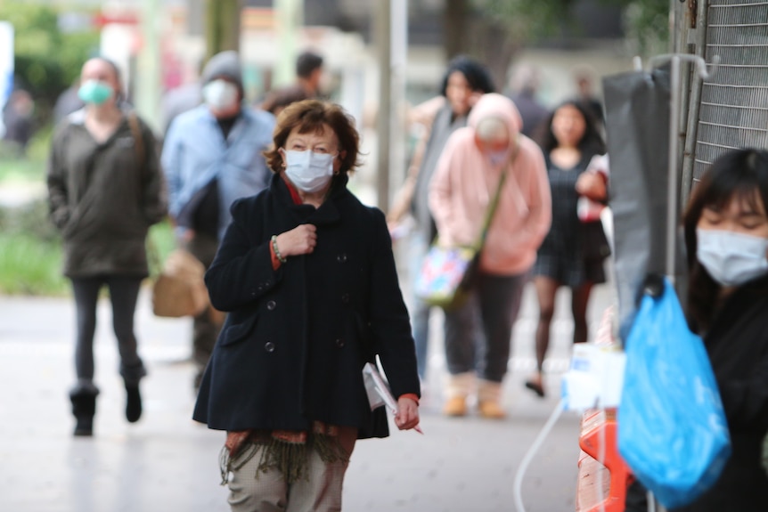 People of all ages are walking outdoors wearing blue surgical masks.