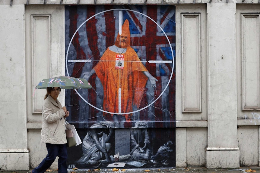 A woman with an umbrella walks past street art of the Union Jack and Assange in an orange case 'free press' lanyard