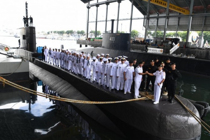 From above, you view two rows of navy sailors standing on the top of a partially-submerged submarine.