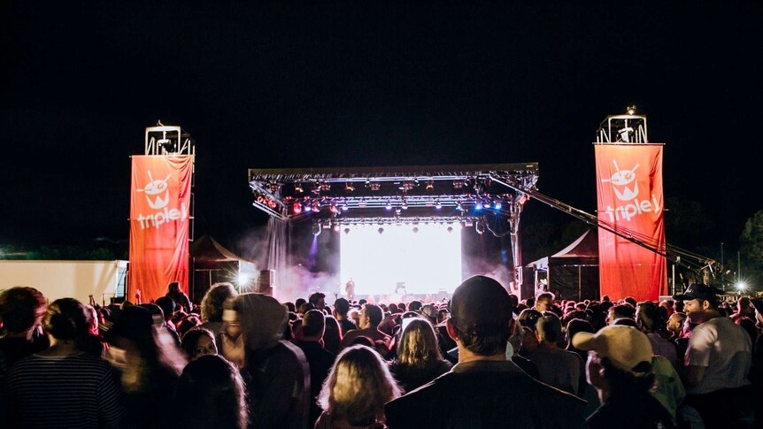 A crowd stands outside looking toward a large stage which is lit up at night and mounted with triple j banners.