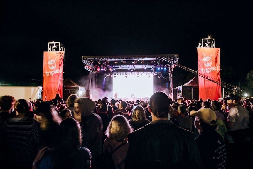 A crowd stands outside looking toward a large stage which is lit up at night and mounted with triple j banners.