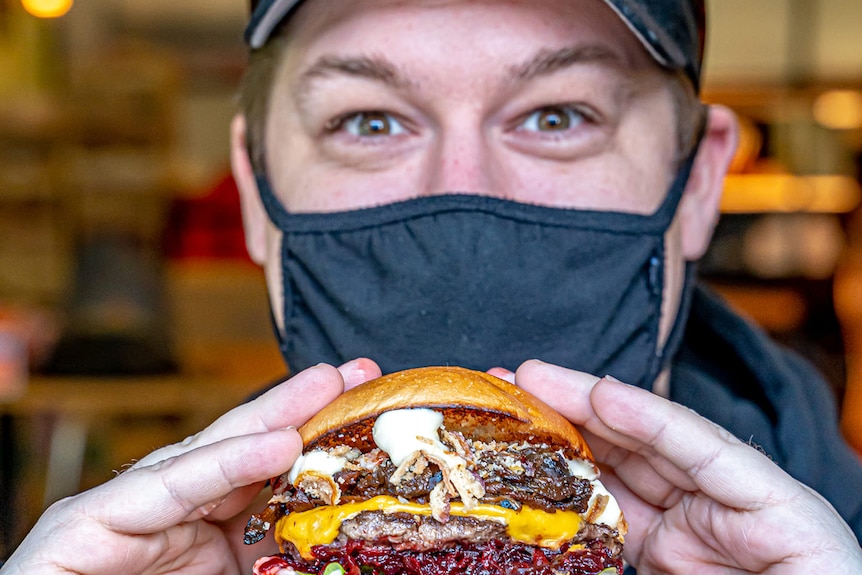 Lachlan Stevens holds a burger out to the camera while wearing a black mask.