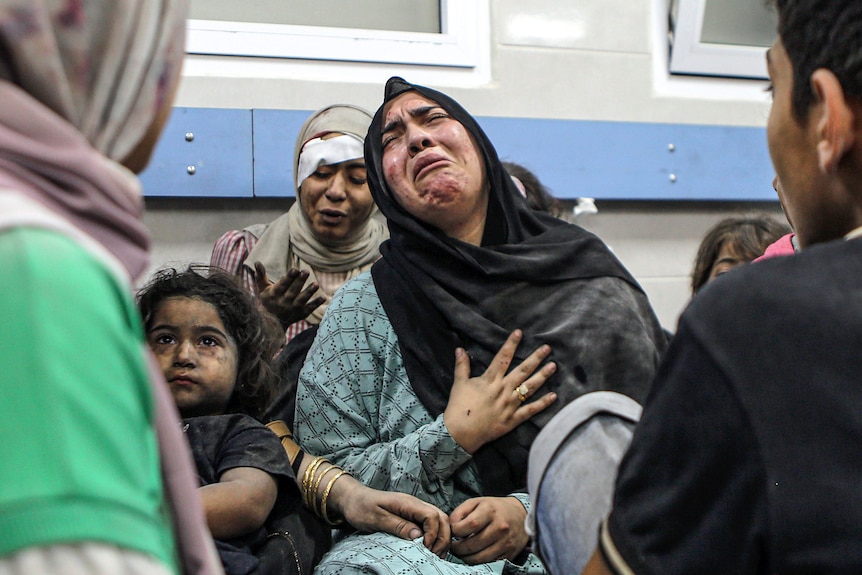 A woman cries as she sits next to a girl covered in dust and in front of an injured woman with an eye bandage.