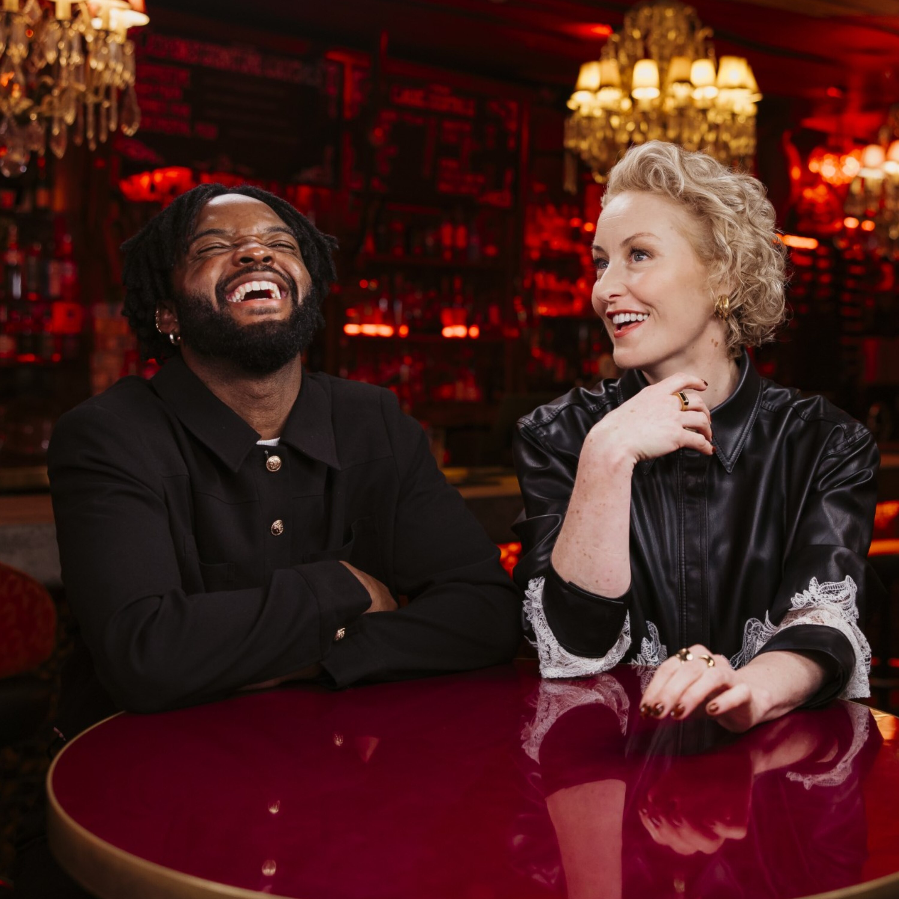 Genesis Owusu and Zan Rowe sitting in a red-tinged bar, laughing