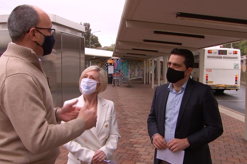 Two men with face masks talk to a woman at a bus station